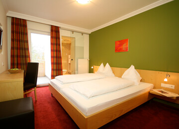 Gemütliches Hotelzimmer mit Doppelbett, grün gestrichener Wand, Schreibtisch und Zugang zu einem Balkon.