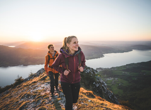 Zwei Wanderer genießen den Sonnenuntergang auf einem Bergpfad mit Blick auf den See und die Landschaft.