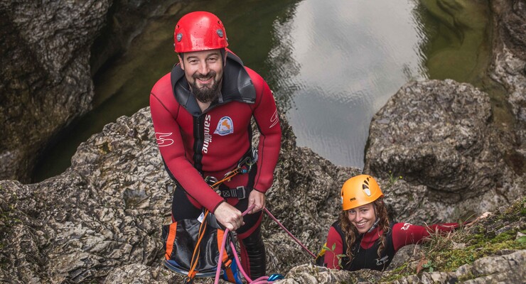 Zwei Personen in Canyoningausrüstung klettern auf felsigem Gelände. Sie tragen Helme und Neoprenanzüge, mit einem Wasserbecken im Hintergrund.