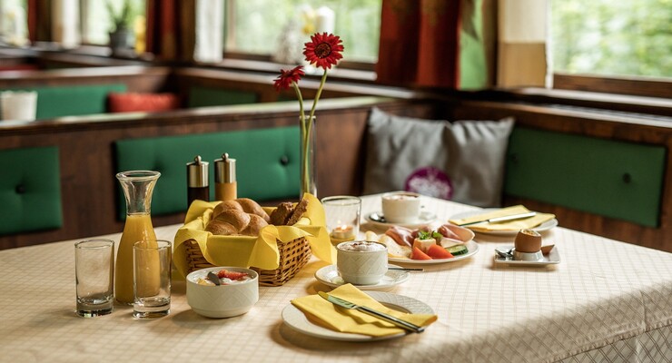 Frühstückstisch in einem gemütlichen Restaurant, gedeckt mit Brötchen, Saft, Kaffee, Aufschnitt und Eiern, dekoriert mit roten Blumen.