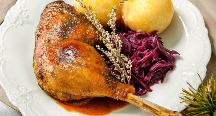 Gericht mit gebratener Gans, Rotkohl und Knödeln auf einem Teller, mit Soßenschüssel und Kerzenlicht auf einem weihnachtlich gedeckten Tisch.