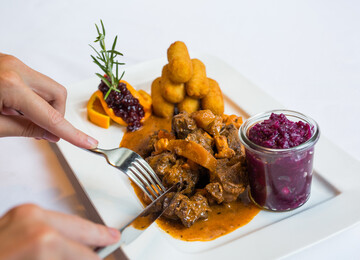 Ein Teller mit einem Fleischgericht, Kroketten, Preiselbeeren und Rotkraut in einem Glas. Hände schneiden das Fleisch.