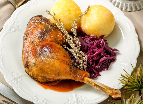 Gericht mit gebratener Gans, Rotkohl und Knödeln auf einem Teller, mit Soßenschüssel und Kerzenlicht auf einem weihnachtlich gedeckten Tisch.