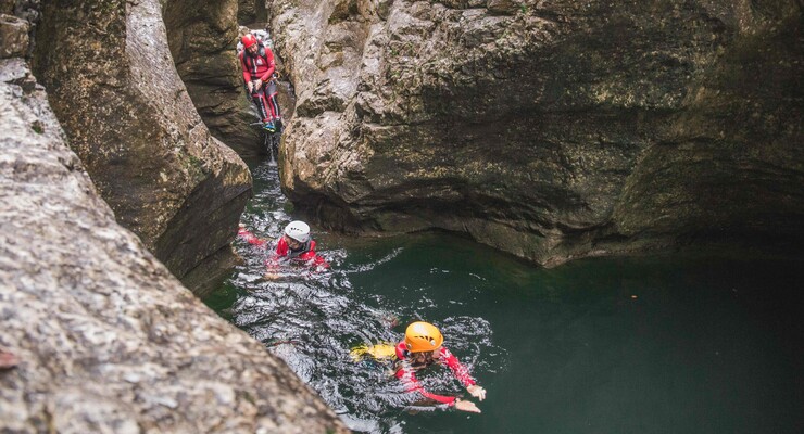 Personen beim Canyoning in einer felsigen Schlucht, ausgerüstet mit Helmen und Neoprenanzügen, schwimmen durch klares Wasser.