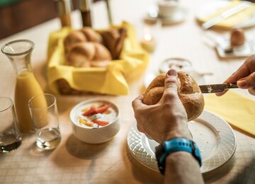 Ein gedeckter Frühstückstisch mit Semmeln, Orangensaft und Joghurt mit Früchten. Hände schneiden eine Semmel auf.