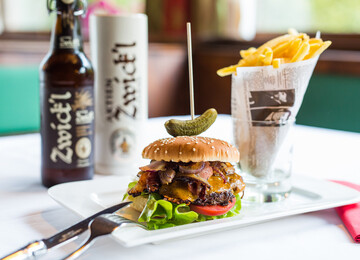 Burger mit Speck und Käse, garniert mit Essiggurke, auf einem Teller. Pommes frites in Papier serviert. Flasche und Glas Zwickl-Bier im Hintergrund.