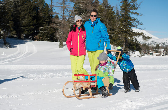 Eine Familie beim Rodeln im Schnee vor einer Winterlandschaft.