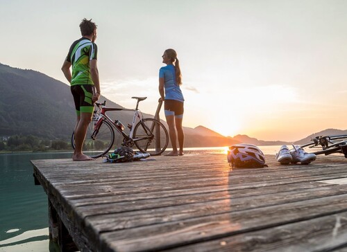 Zwei Radfahrer stehen auf einem Steg am See bei Sonnenuntergang, mit Fahrrädern und Helmen auf dem Holz.
