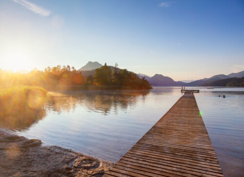Ein Holzsteg erstreckt sich über einen ruhigen See in der Morgensonne, umgeben von Bergen und Bäumen.