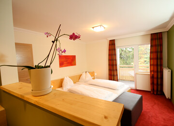 Gemütliches Hotelzimmer mit Doppelbett, rotem Teppich und Balkonblick. Eine Orchidee steht auf der Holzablage im Vordergrund.