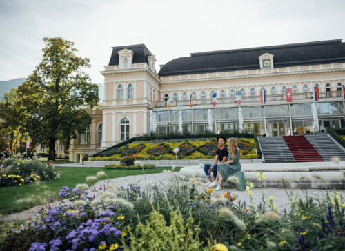 Ein Paar sitzt auf einer Bank vor dem Kongresshaus in Bad Ischl mit bunten Blumen im Vordergrund und mehreren Flaggen auf dem Balkon.