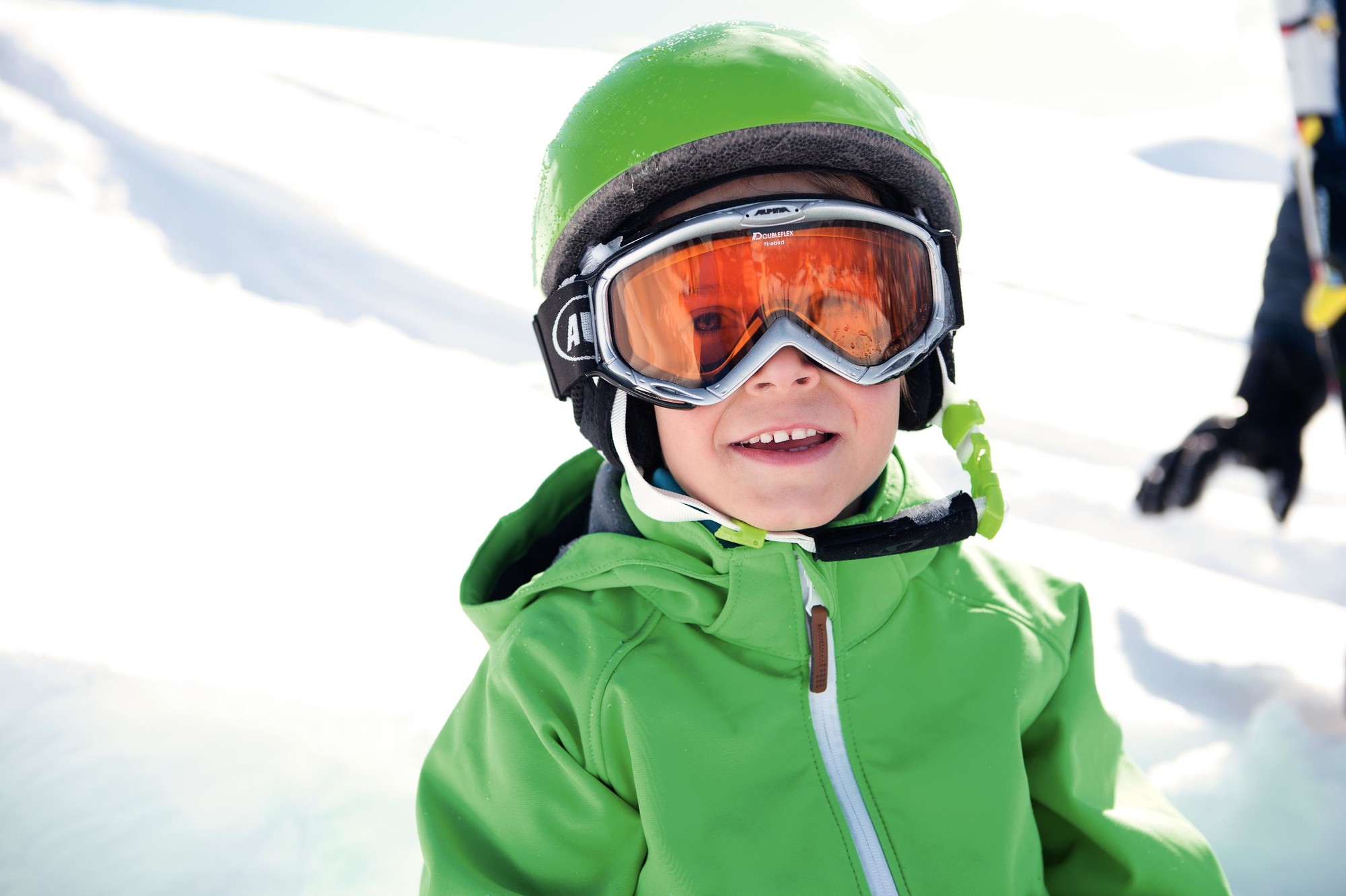 Kind auf Skipiste in grüner Jacke und Helm, lächelnd in die Kamera.