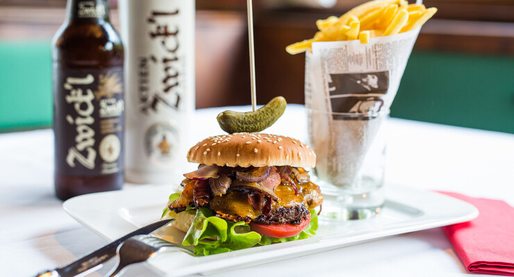Burger mit Speck und Käse, garniert mit Essiggurke, auf einem Teller. Pommes frites in Papier serviert. Flasche und Glas Zwickl-Bier im Hintergrund.