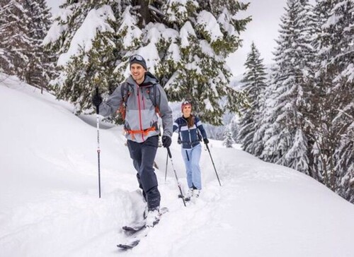 Zwei Personen beim Skitourengehen auf verschneitem Waldweg, umgeben von schneebedeckten Bäumen.