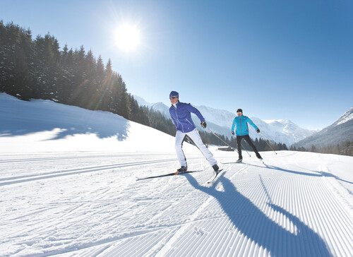 Zwei Personen beim Skilanglauf auf einer sonnigen, verschneiten Landschaft mit Bergen im Hintergrund.