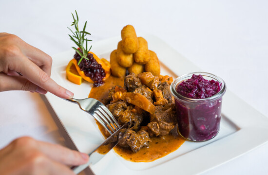 Ein Teller mit einem Fleischgericht, Kroketten, Preiselbeeren und Rotkraut in einem Glas. Hände schneiden das Fleisch.